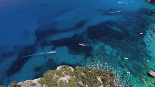 Top-down view of two boats crossing turquoise water and dark seagrass patches as an FPV drone flies down the rugged Cala Goloritze coast, Baunei, Sardinia.