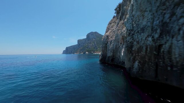 A sweeping reveal shot of Cala Goloritze bay, Baunei coast, Sardinia &mdash; FPV drone banking left toward open sea, boats anchored in turquoise water below.