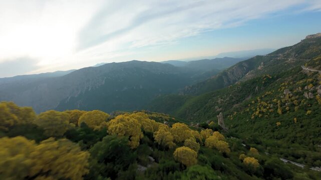 Cinematic FPV drone footage flying in close proximity to a towering rock pillar, then along a mountain road with passing cars, hillsides covered in blooming yellow Mount Etna broom, Baunei, Sardinia.