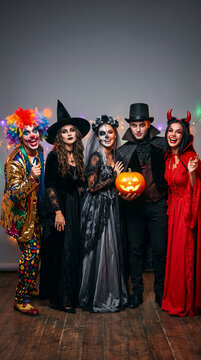 Group of people in Halloween costumes like a clown, witch, bride of death, vampire, and devil, celebrating the holiday with a jack-o-lantern