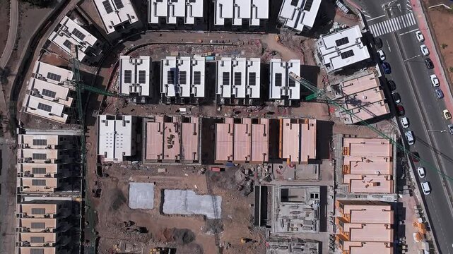 Top-down drone view of a residential construction site with rows of modern houses, unfinished rooftops, solar panels and surrounding roads, showing different stages of urban development