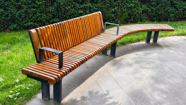Curved wooden park bench with black metal armrests and legs offering comfortable outdoor seating in a public garden or green space, representing relaxation and urban design