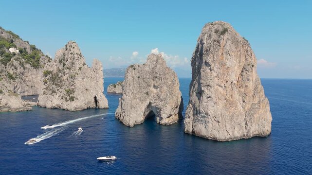 Boats are seen gliding through the clear blue water near the large rocks on the coast of Capri, Italy. The sun shines brightly over the setting, creating a calm atmosphere.