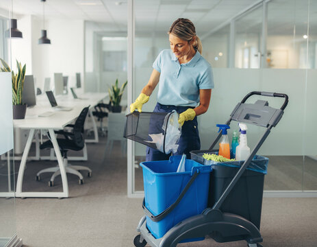 A female cleaner in yellow gloves emptying a trash bin into a plastic bucket on a janitor trolley in a modern office.