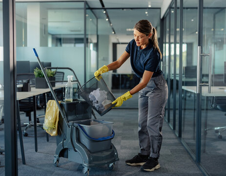 A female cleaner in yellow gloves emptying a trash bin into a plastic bucket on a janitor trolley in a modern office.