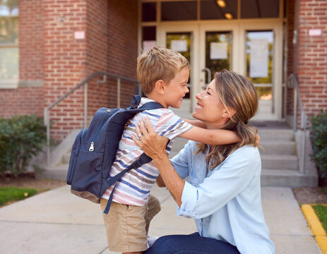 A mother hugging her son outside a school building. A happy mom embraces her boy with his backpack, depicting a back-to-school or reunion concept.