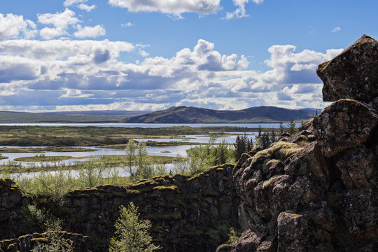 Wetlands and lake lie between dark rift rocks in Thingvellir National Park, Iceland. Green channels extend toward blue water and distant mountains. Basalt edges border open lowlands.