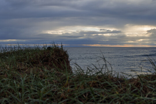 A grassy beach faces an Atlantic cove at Stora Sandvik on the Reykjanes Peninsula, Iceland. Soft sunset light reaches layered dunes, pale surf, black cliffs, and a glowing break in storm clouds.