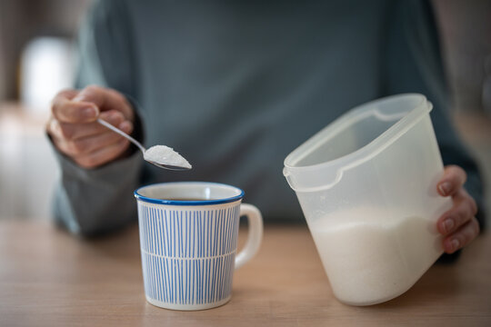 Woman holds spoonful of white sugar over coffee beside sugar container, showing habitual sweetening, sugar dependence and difficulty reducing added sugar despite concerns and efforts to quit. 