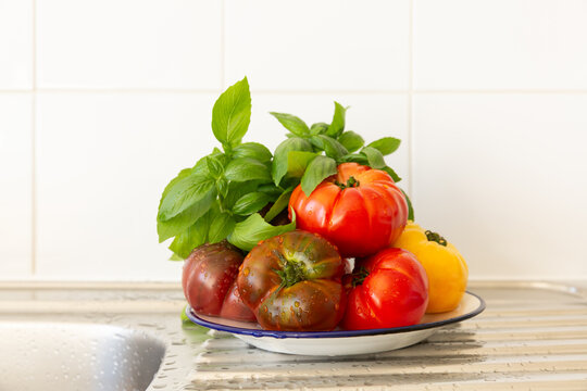 Assiette avec des tomates rouges et jaunes empil&eacute;es et un bouquet de basilic frais pos&eacute;e sur un &eacute;vier de cuisine en inox, ensemble de l&eacute;gumes frais et crus