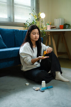 Young Asian Woman Sitting on Floor Holding Pills with Medication Scattered Around Her at Home