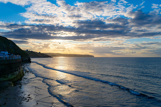 vening sun breaking through dramatic clouds over the North Sea, with gentle waves reaching the shore beside Whitby Harbour in North Yorkshire, England.