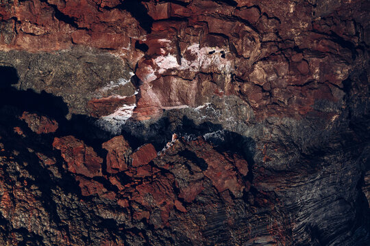 Lava forms diagonal ridges, cracks, and porous hollows inside Raufarholshellir, Iceland. Iron-red tones cross the broad surface as subdued cave light reveals relief and shadowed gaps below.