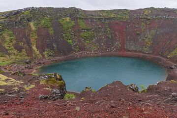 A turquoise lake fills the oval basin of Kerid crater in Iceland. From the wet rim, blue water sits below layered red scoria walls, revealing the full volcanic bowl beneath low cloud. © Artem