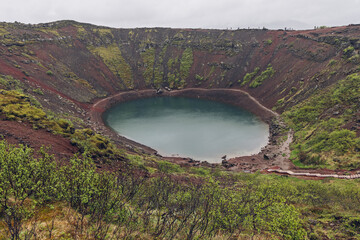 A rim path circles red walls and turquoise water at Kerid Crater, Iceland. Diffuse afternoon light illuminates wet scoria and moss above the crater lake. The curved trail echoes the lake outline. © Artem