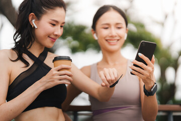 Two Asian women in athletic wear using smartphone and smartwatch outdoors