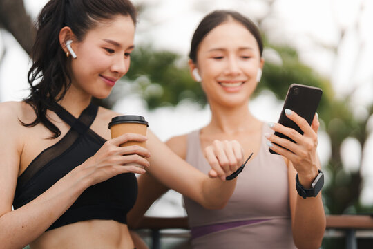 Two Asian women in athletic wear using smartphone and smartwatch outdoors