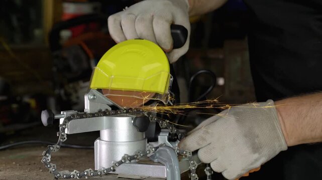 Worker in gray gloves operates a chain sharpening tool with a yellow guard, sparks flying as the chain is sharpened, showcasing a workshop environment filled with tools and equipment