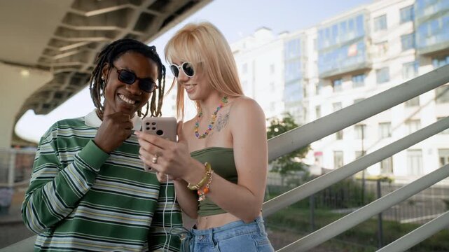 African American young man and Caucasian young woman standing outdoors, smiling and looking at smartphone screen together. Footage is ideal for mobile app marketing or social media campaigns