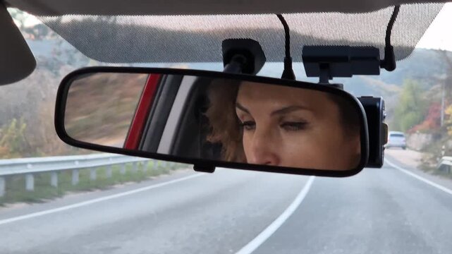 Woman driving, focus on reflection in the rearview mirror while traveling along a scenic winding highway road. Interior steady shot.