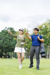 Obraz premium Two smiling Asian golf players walking on a green course, carrying clubs and a bag, pointing and chatting as they plan strategy and enjoy a sunny day outdoors