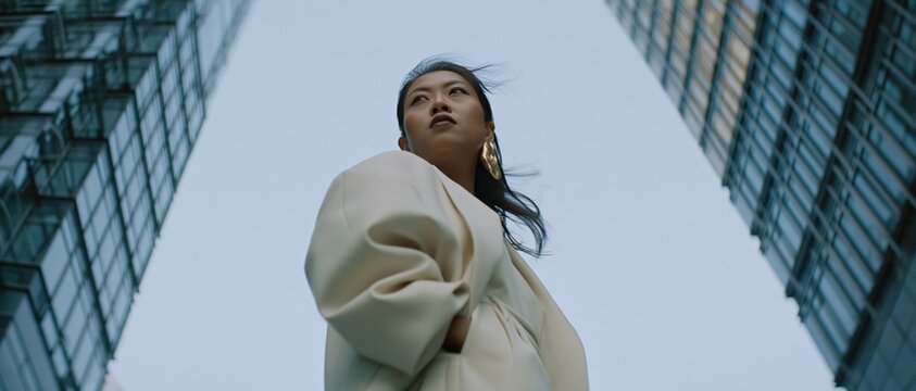 Confident Asian woman looking up between towering city skyscrapers, symbolizing ambition and urban success