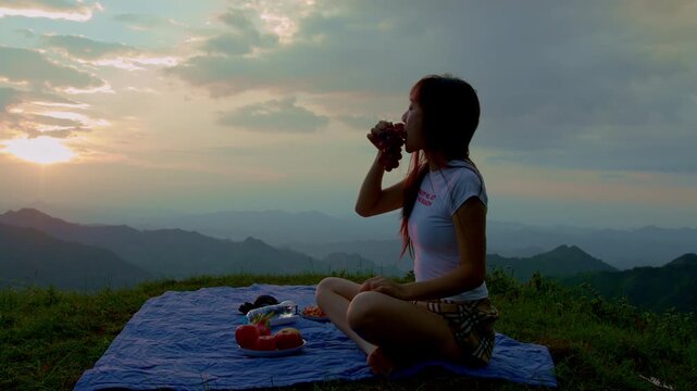 Woman enjoying grapefruit while sitting on a picnic blanket at sunset, surrounded by layered mountain ridges and warm evening light in a peaceful outdoor travel and lifestyle scene