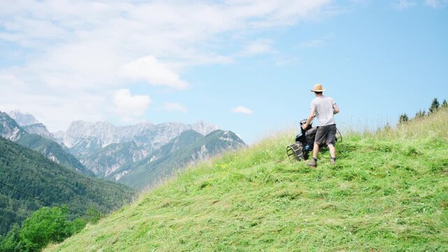 A farmer works on a small alpine farm, mowing grass on a hillside. The bright sky and distant mountains create a backdrop for this daily task on a private property.