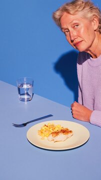 Woman in her fifties looking at a small portion of food on a plate, featuring a surreal oversized shadow on a blue surface with copy space