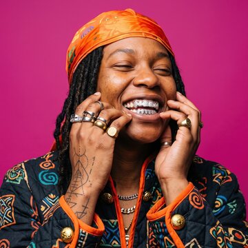 Stylish Black person with silver grillz and orange headscarf, happy individual with dreadlocks and gold rings, vibrant pink background portrait
