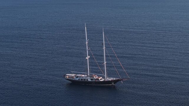 Elevated aerial view of a two-masted sailing vessel anchored alone on calm blue water, surrounded by expansive open sea and uncluttered copy space.