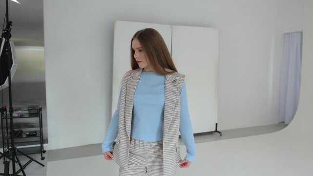 Beautiful young female model wearing a stylish plaid suit posing and dancing playfully in a professional photo studio with a white cyclorama and lighting equipment in the background