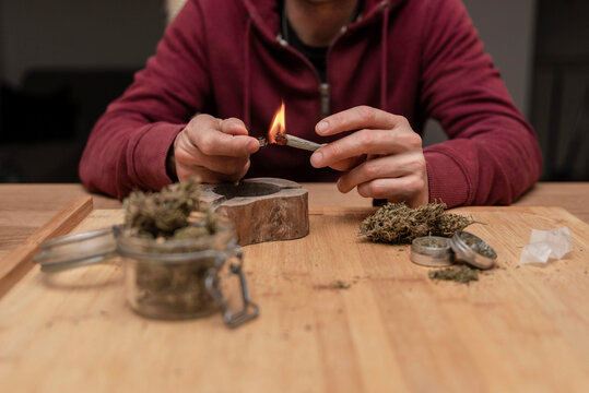 Anonymous man lighting blunt while smoking cannabis sitting at wooden table with marijuana in glass jar