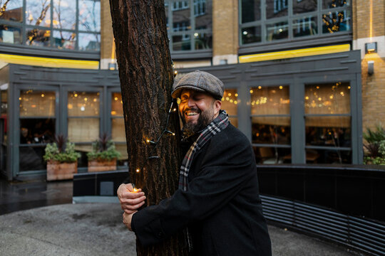A smiling man hugs a decorated tree at an outdoor winter market