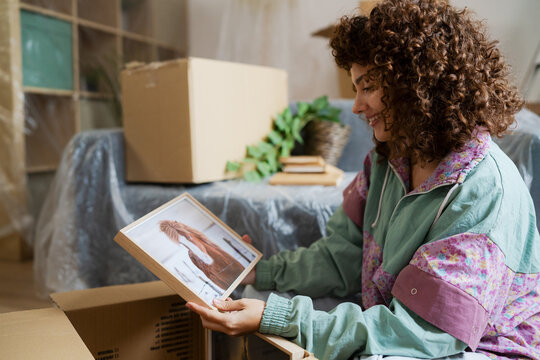 Smiling woman unpacking box and holding framed photo in new home