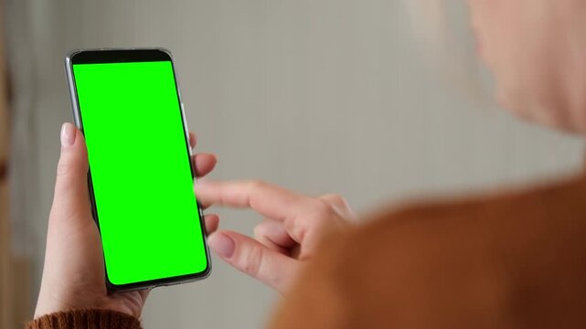 Over the shoulder view of woman holding smartphone vertically and touching green screen mockup chroma key display. Girl tapping on mobile phone template mockup blank screen inside a cozy room