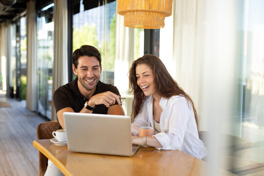 Happy couple laughs while sharing a laptop at a cafe table