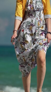 Summer vacation fashion, a young woman walks along the beach with floral dress blowing in breeze. Vertical steady low-angle tracking shot.