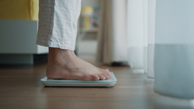 A person in white pajamas standing barefoot on a digital weight scale placed on a wooden floor near a brightly lit window