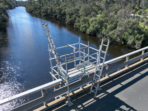 Scaffolding tower installed on a rural bridge to provide safe access for maintenance and inspection work. Metal platforms and supports surround the bridge structure over a small waterway.