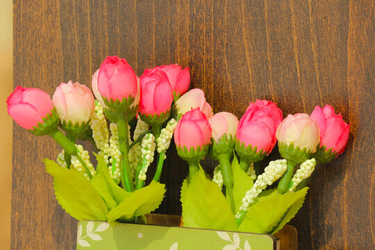 Pink rose buds in wall planter on dark wood background, decorative artificial flowers bouquet with green leaves on wooden texture panel for romantic home interior decor