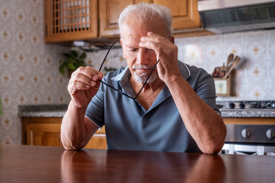Senior man suffering from a severe headache or migraine, feeling stressed, tired, and worried about his health while holding his glasses in a home kitchen setting
