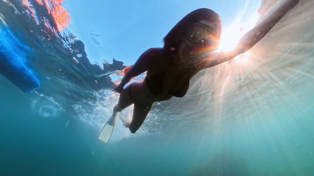 Crystal clear sea, a woman wearing fins glides gracefully near the ocean surface with sunlight beaming down. Tracking shot.