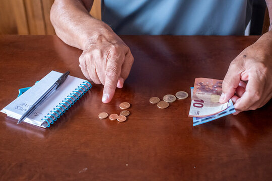 Senior person's hands counting euro coins and banknotes on a wooden table next to a notebook with savings home written, focusing on financial planning and retirement budgeting
