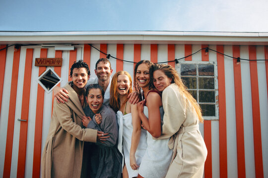 Friends enjoy wellness and sauna community outside Nordic bathhouse in robes and towels