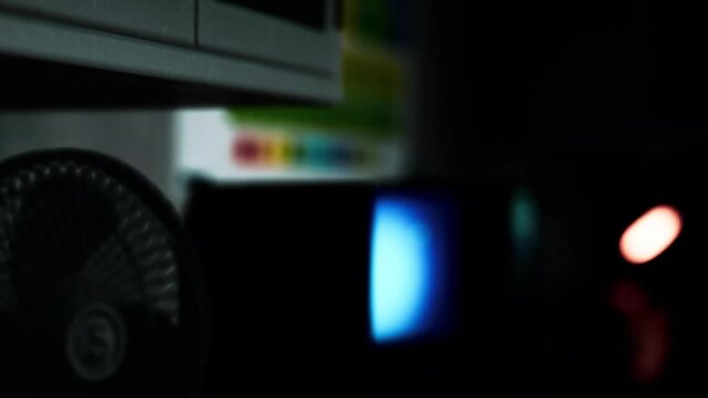 In a dark room, multiple electronic devices like screens and a fan are visible. Soft light from the screens creates a contrast with the darkness. The atmosphere feels mysterious and tech focused.