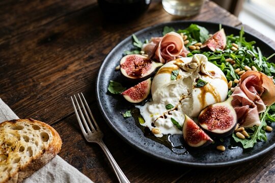 Fresh Italian burrata cheese torn open with ripe figs, prosciutto, baby arugula, pine nuts, and balsamic glaze on artisanal plate