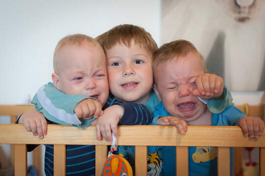 Elder brother hugging his two crying baby twin siblings inside a wooden crib during a bright morning at home while providing comforting love