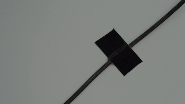 Closeup hands applying small tape to cable on gray surface, careful peel and stick motion, precise alignment of black square patch to secure wire and prevent tripping