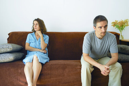 Upset man and woman sitting at opposite ends of brown sofa looking away. Represents emotional distance after argument, relationship silent treatment, and communication breakdown between partners.